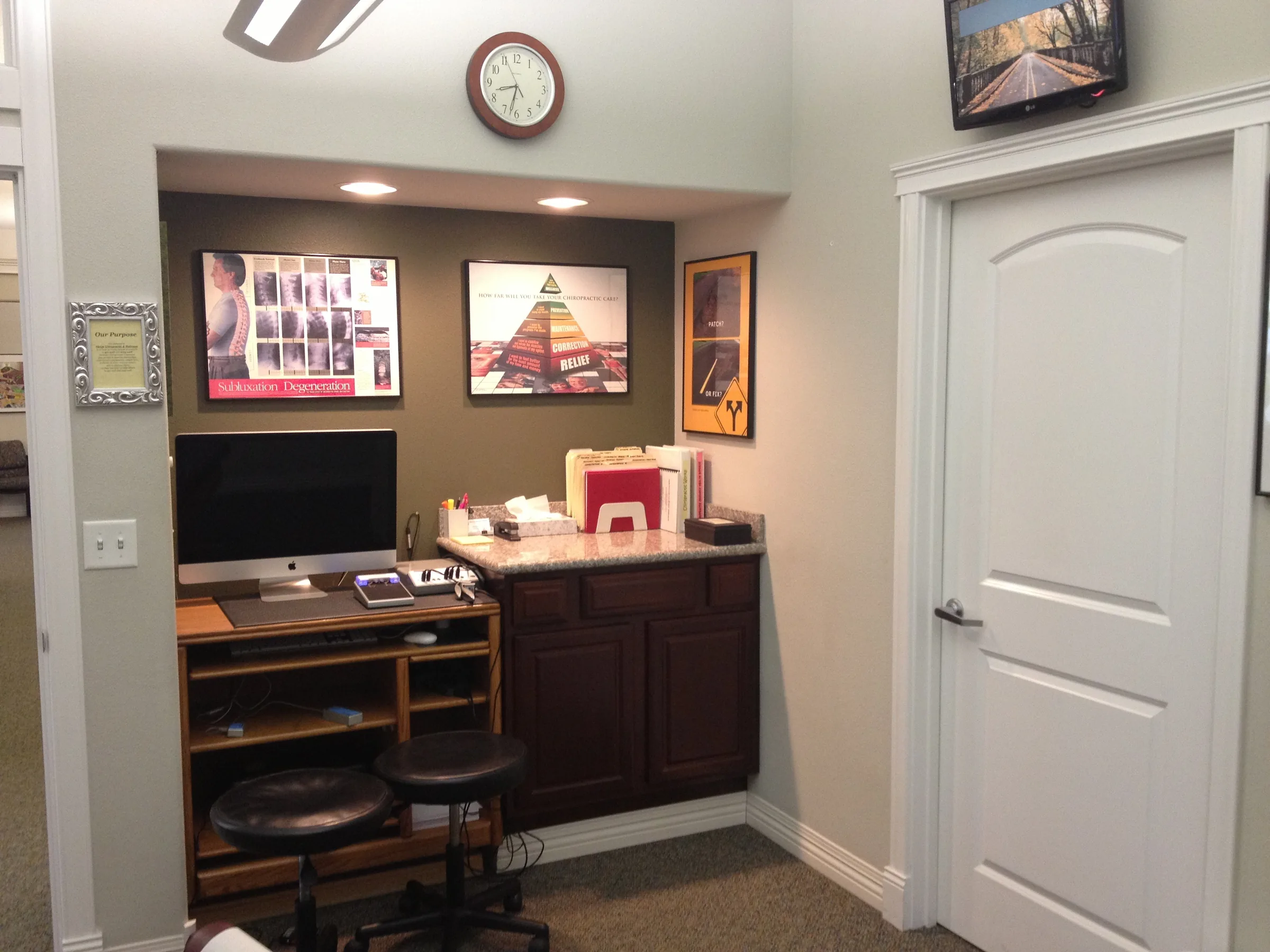 Chiropractic office consultation room with desk, computer, educational posters, and clock on wall.