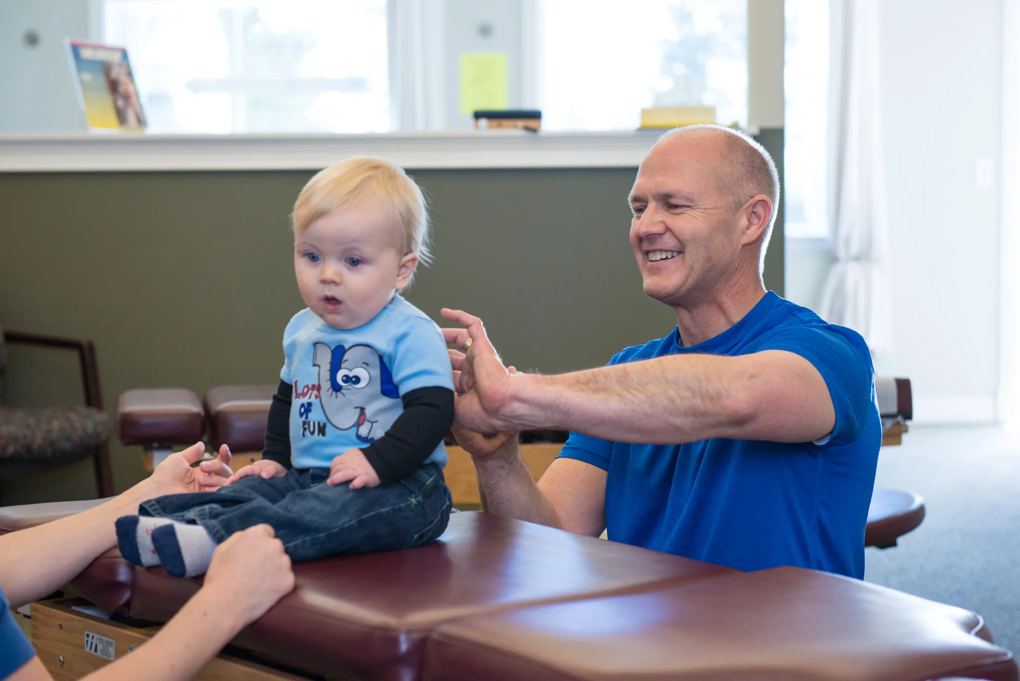 Chiropractor in blue shirt examining a toddler at a table in a bright office.